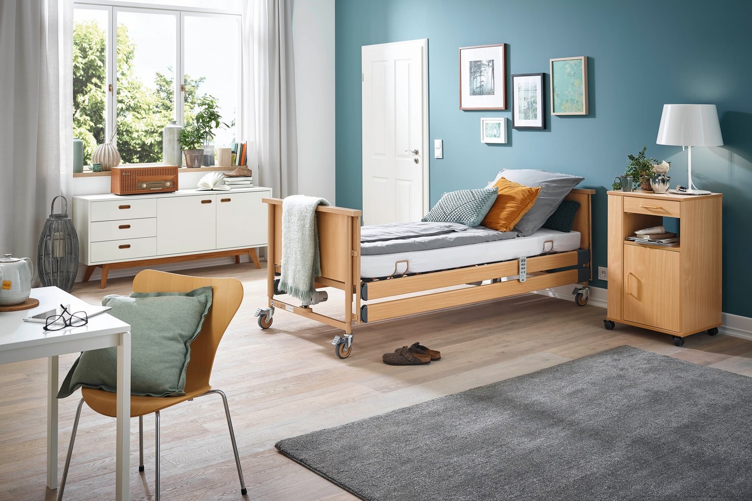 Stilvoll eingerichtetes Pflegezimmer mit höhenverstellbarem Pflegebett, gemütlicher Sitzecke und großem Fenster