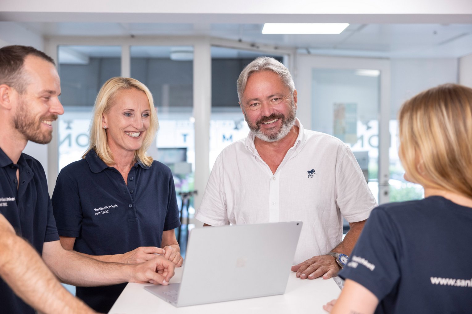 Vier MOT-Mitarbeitende stehen zusammen um einen Tisch und besprechen etwas am Laptop in freundlicher Atmosphäre