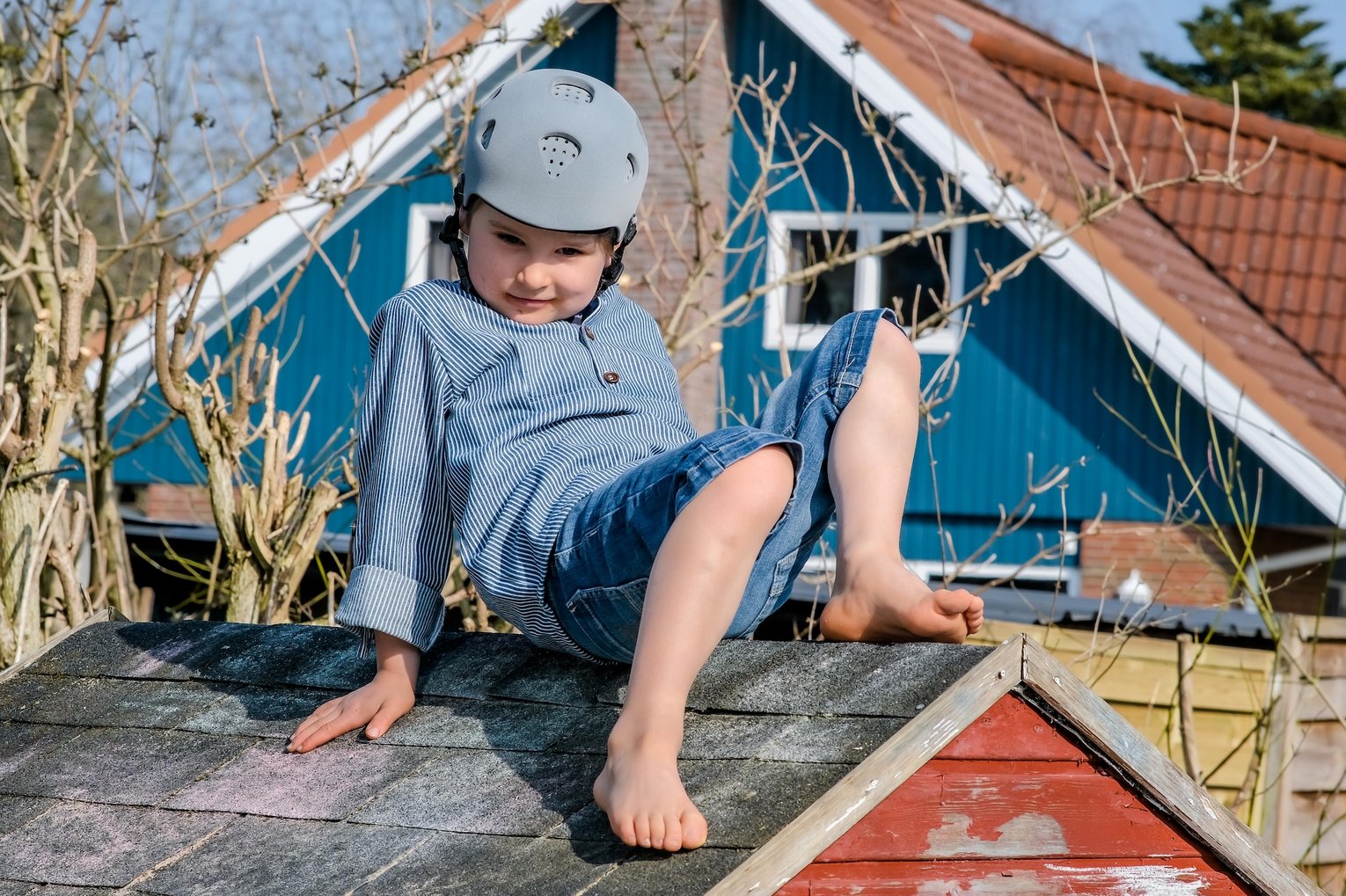 Kind mit grauem Schutzhelm und Kapuzenpullover sitzt barfuß auf einem Holzhausdach und spielt draußen